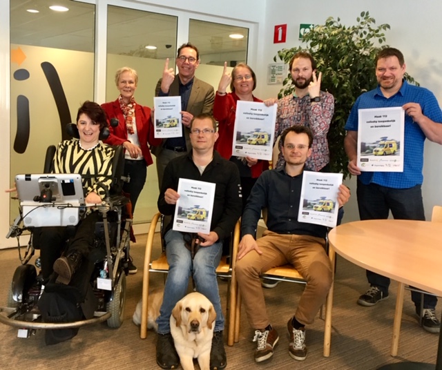 Leden van de 112-Alliantie tonen trots hun position paper. Zittend op voorste rij: een mevrouw in een rolstoel, een meneer met een geleidehond, een meneer die het position paper toont. Daarachter staan twee vrouwen en drie mannen. De drie middelste personen gebaren 1 – 1 – 2.