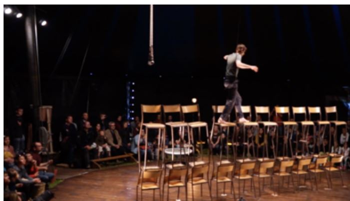 Publiek kijkt naar een circusartiest die zich over een gebouwde toren van stoelen maneuvreert