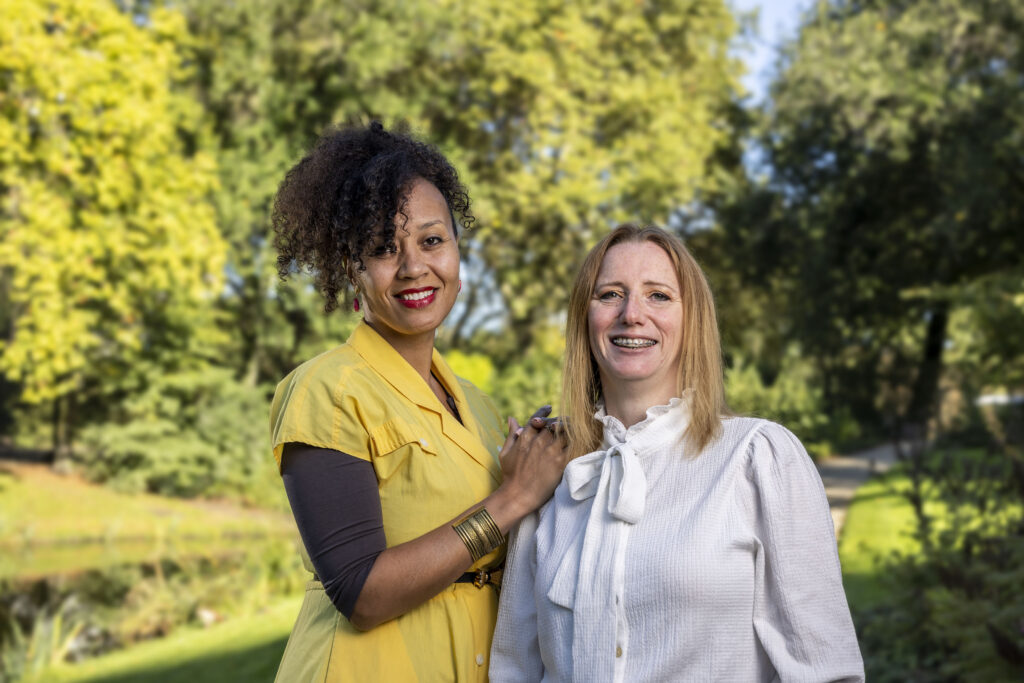 Vrouw met gele jurk en vrouw met witte blouse kijken vriendelijk naar de camera