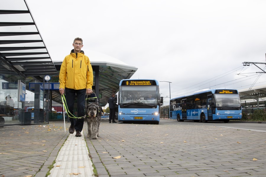 Man met stok en hond loopt over geleidelijn bij busstations