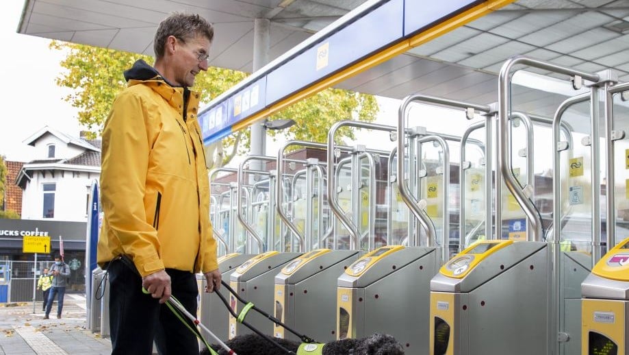 Man met gele jas stok en hond loopt richting poortjes op station