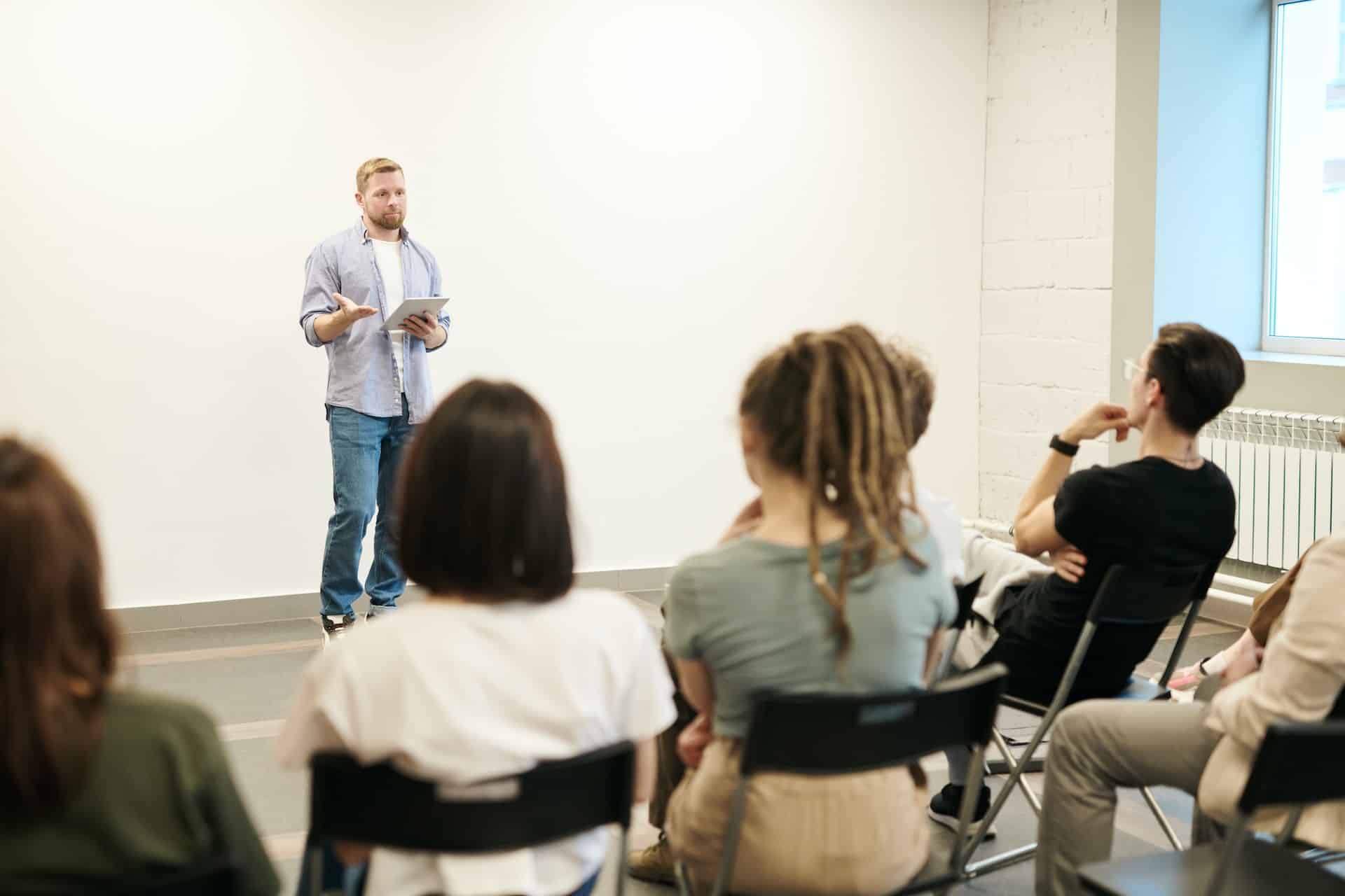 Een man staat voor een zittend publiek in een witte lesruimte en geeft een training.