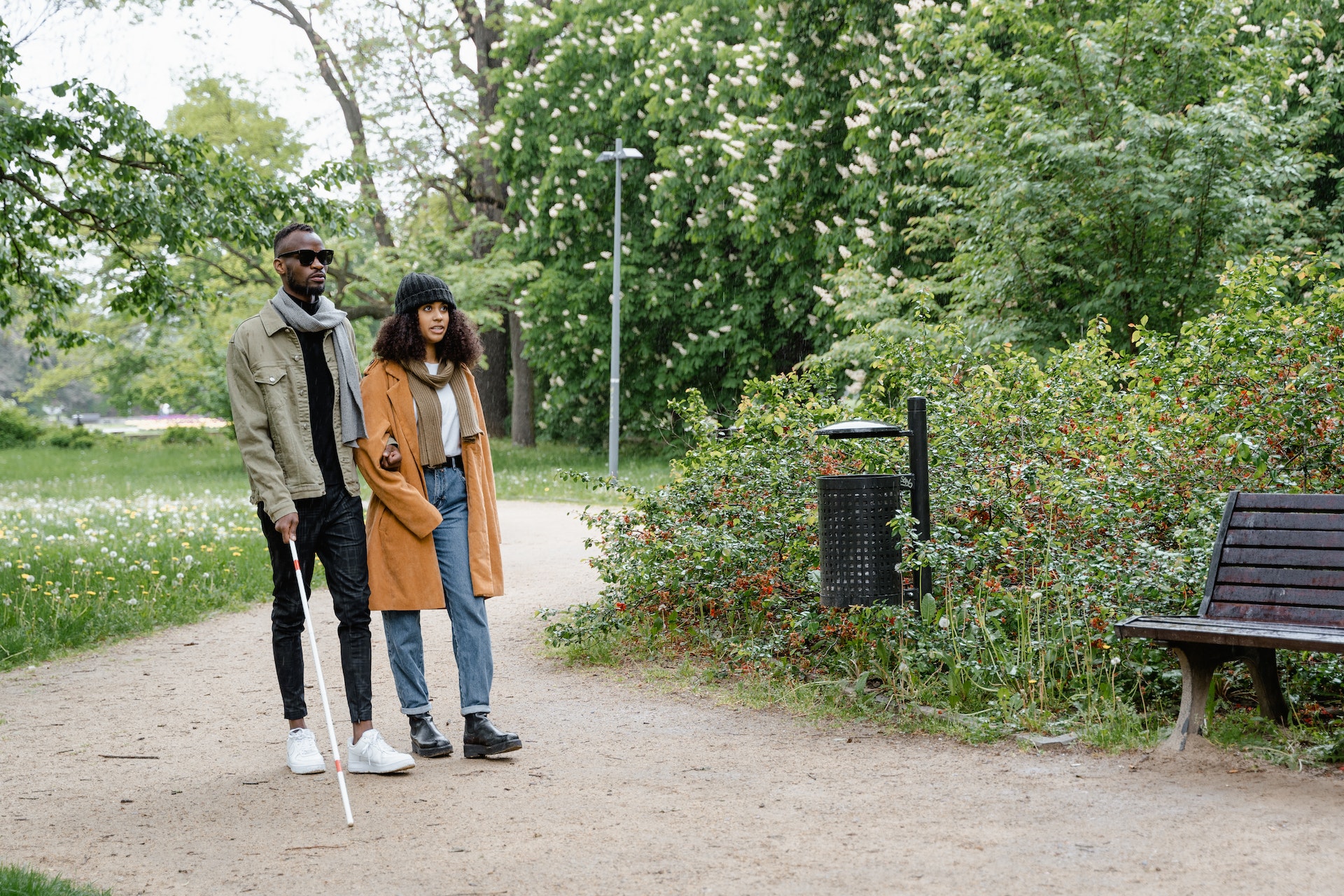 Een blinde man met geleidestok loopt arm in arm met een vrouw in het park.