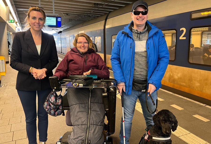 Staatssecretaris met Peter Waalboer en zijn hond en Karin van Dijk in een elektrische rolstoel poseren voor een trein