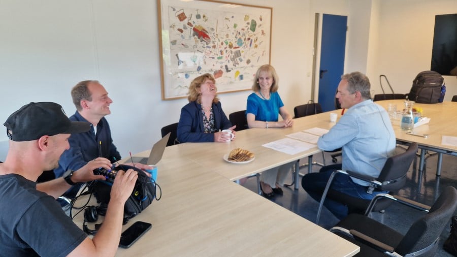 Twee sprekers, de leider van de podcast en mensen van de techniek zitten aan tafel