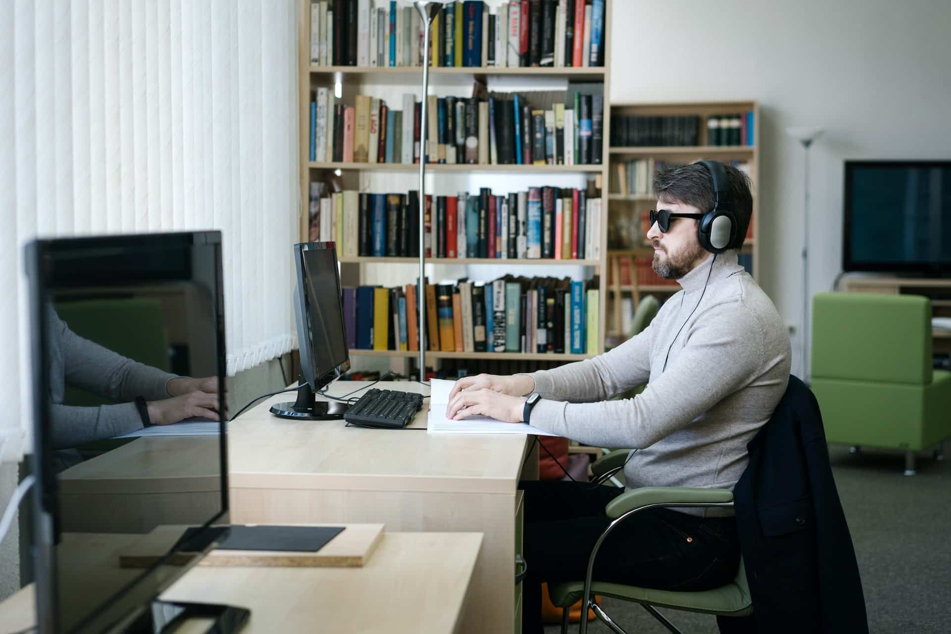 Een blinde man met een zonnebril en koptelefoon op zit achter een computer te lezen.