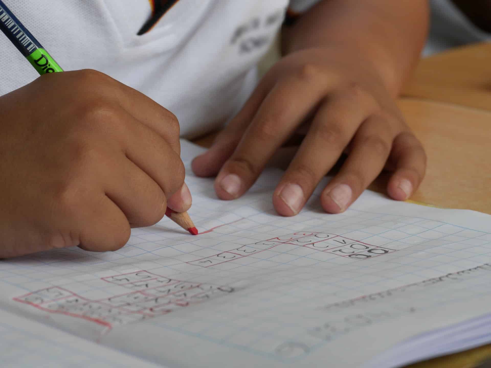 Twee kinderhanden tekenen wiskundige tabellen met een rood kleurpotlood in een schrift.