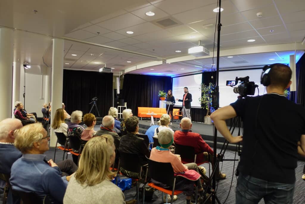 een volle zaal met toeschouwers kijkt naar twee sprekers op een podium die worden gefilmd door een camera