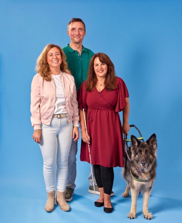 Een man en twee vrouwen, waarvan één met geleidehond en taststok