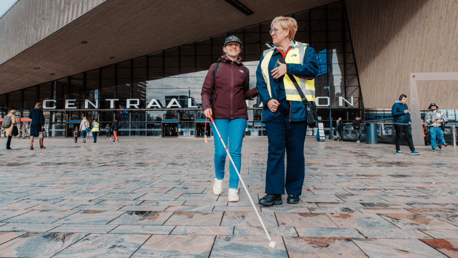 een jongeman met taststok krijgt hulp van een reisassistent op het plein voor rotterdam centraal