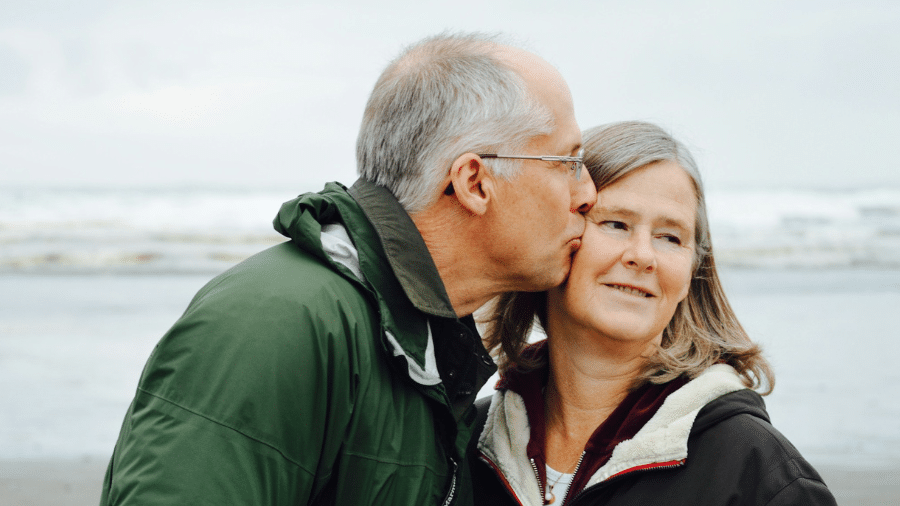 een man kust een vrouw op haar wang op een strand, de lucht is lichtelijk grauw
