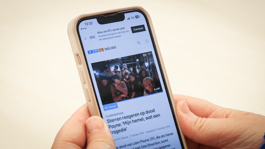 twee handen houden een smartphone vast, in beeld staat een nieuwsartikel van RTL Nieuws met de kop: Sterren reageren op dood Payne: 'Mijn hemel, wat een tragedie'. Onder de kop staat een donkere, drukke persfoto met veel mensen erop. Ze lijken te rouwen. In de achtergrond staan fotografen met hun camera in de lucht over de groep mensen heen te fotograferen.