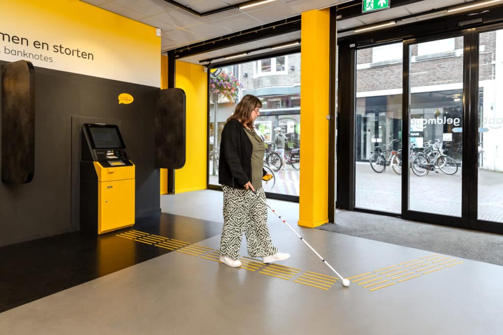 een ervaringsdeskundige navigeert met een witte stok over de geleidelijn richting de uitgang van een winkel, ze heeft zojuist geld gepind.