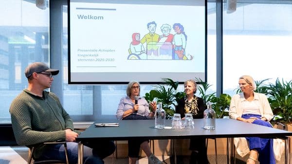 Vier mensen zitten rondom een tafel. Achter hen een scherm met de presentatie van het actieplan toegankelijk stemmen