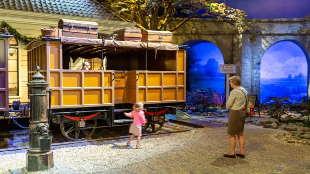 een oude passagierswagon staat op de rails in het spoorwegmuseum. Een jong meisje bekijkt de gele wagon met houte kratten op het afronde dakje. Het vader of opa kijkt van een afstandje toe.
