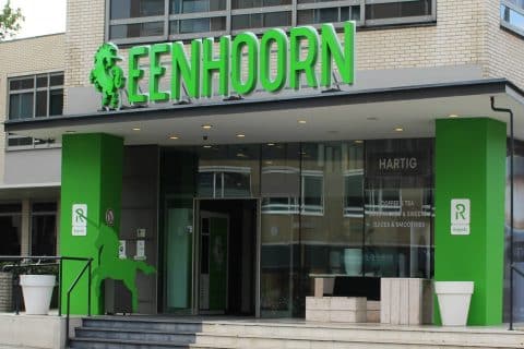 de ingang van meeting centrum de Eenhoorn. E hangt een groot, groen bord met het logo boven de entree. Aan weerszijden van de entree staan grote groene pilaren.