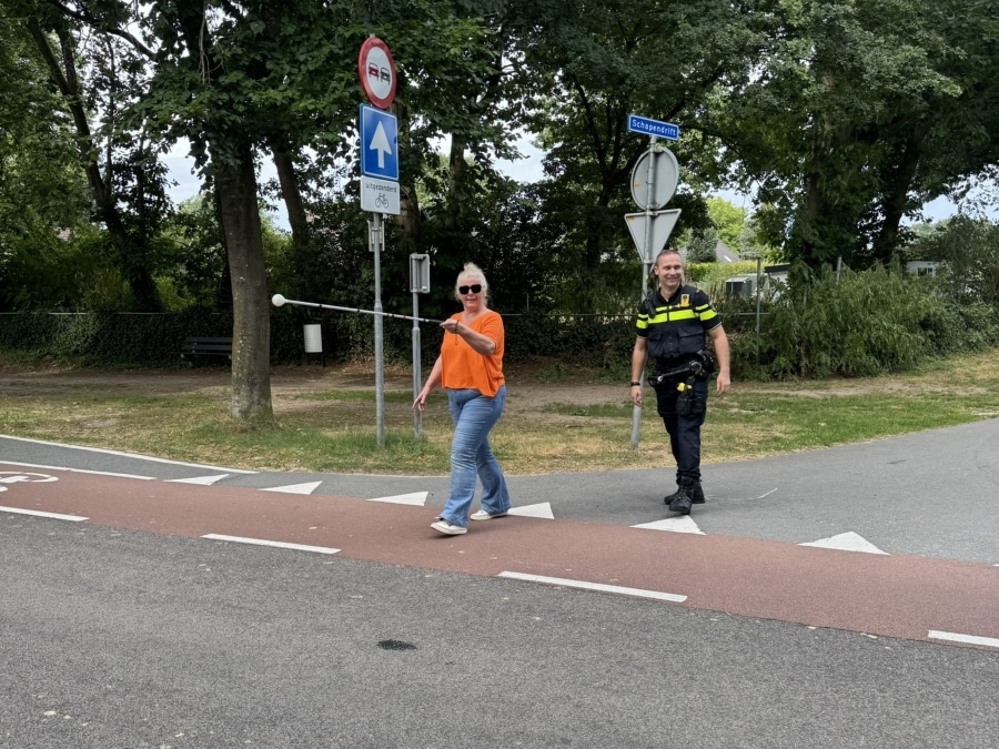 Een vrouw steekt een weg over met de witte stok horizontaal in de lucht. Naast haar loopt een politieagent.