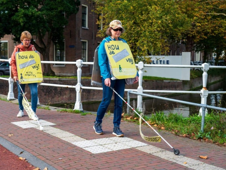 Twee vrouwen lopen met een witte stok over de geleidelijn. Ze dragen een geel bord met de tekst Hou de lijn vrij