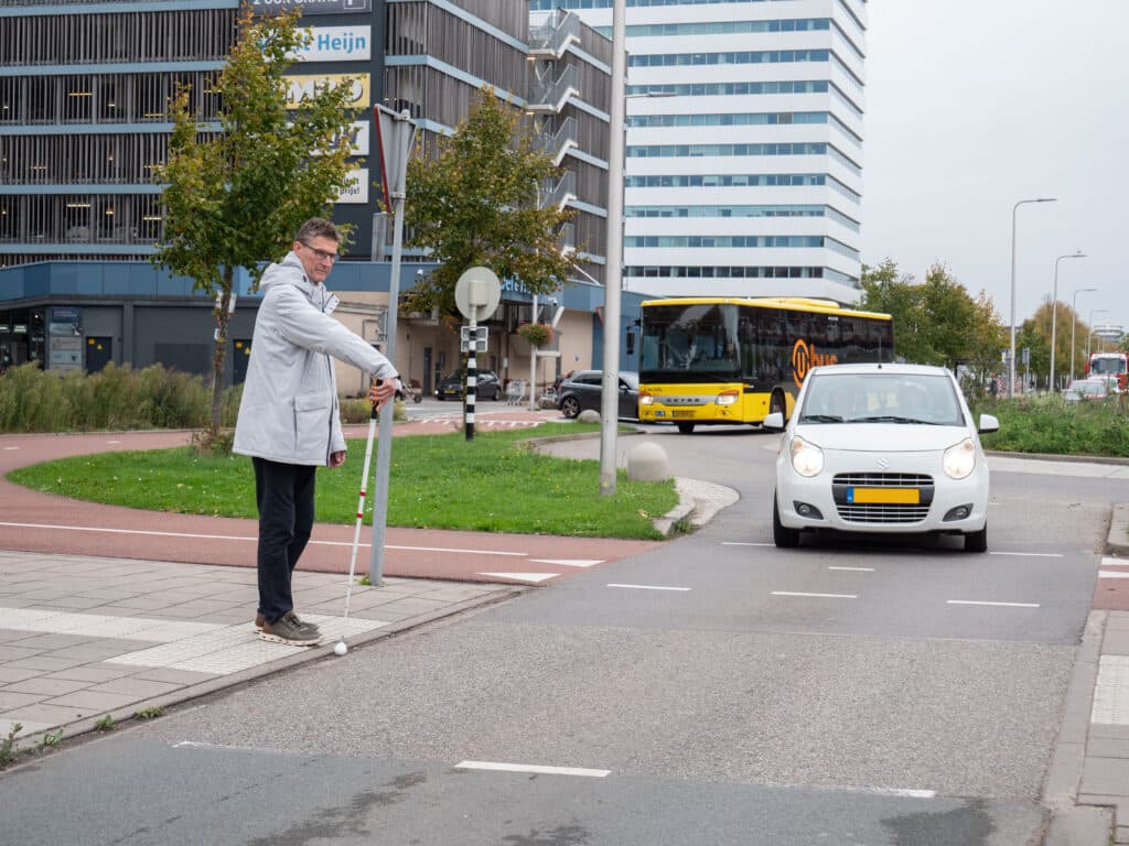 een man met witte stok maakt een stopgebaar, een auto die de rotonde verlaat verleent voorrang