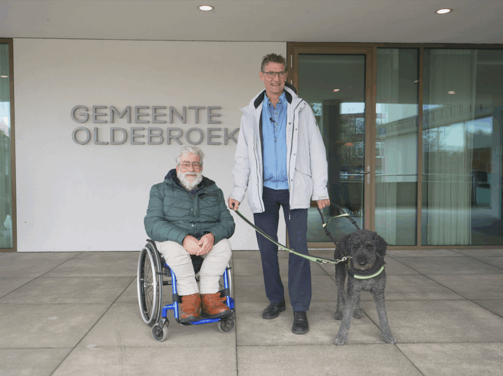 Ton van Weerdenburg met geleidehond Sofie en rolstoelgebruiker Jan Oosterbroek voor de ingang van het gemeentehuis van Oldebroek. De naam van de gemeente staat groot op de witte gevel.