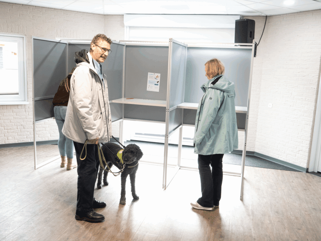 Ton van Weerdenburg met geleidehond Sofie en Hanneke de Boer bekijken een rij stemhokjes.