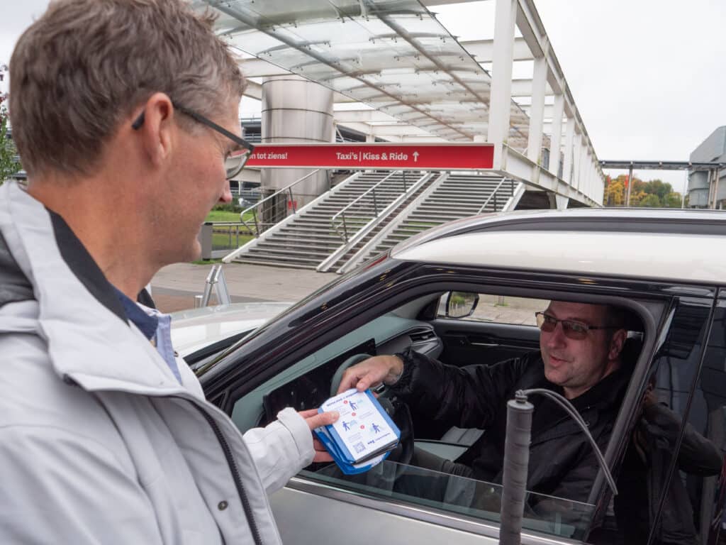 een man met witte stok buigt richting het raam van een auto en rijkt de bestuurder een blauwe parkeerschijf uit