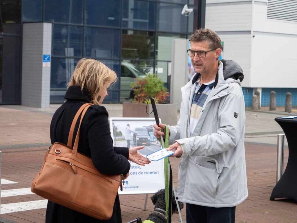 collega Ton van Weerdenburg reikt een vrouw een parkeerschijf uit. Op de achtergrond ligt een zebrapad en staat een groot bord met de tekst: Stop voor de witte stok, geef ons de ruimte!