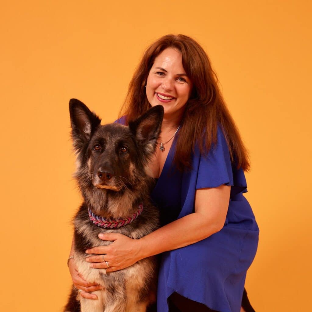 Een vrouw in een blauwe jurk hurkt naast haar geleidehond en kijkt vrolijk in de camera