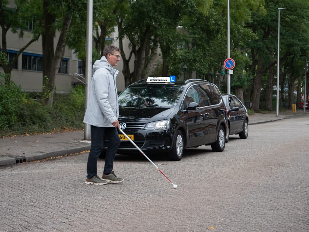een man met witte stok steekt over, een lesauto wacht tot hij aan de overkant is