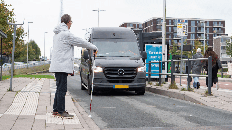 een man met witte stok maakt het oversteekgebaar, een bestelbus komt tot stilstand