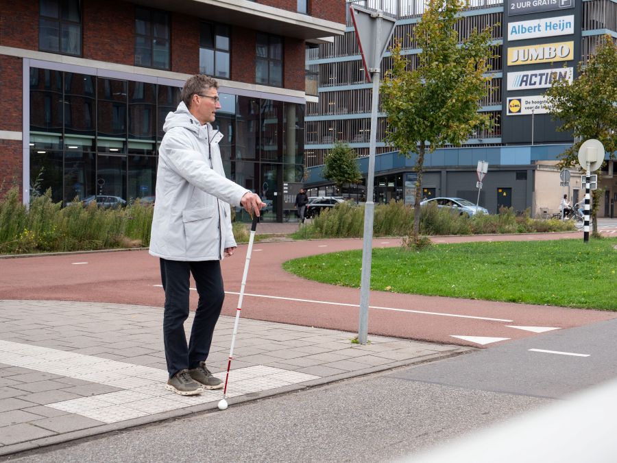 Een man met een witte stok luistert of hij kan oversteken. De stok staat omhoog tegen de stoeprand.