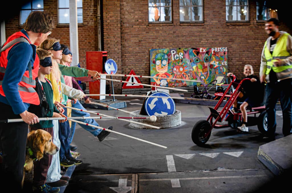 Zeven kinderen met een witte stok en blinddoek steken de weg over in de verkeerstuin in Amersfoort. Een jongen op een skelter wacht geduldig met een grote glimlach.