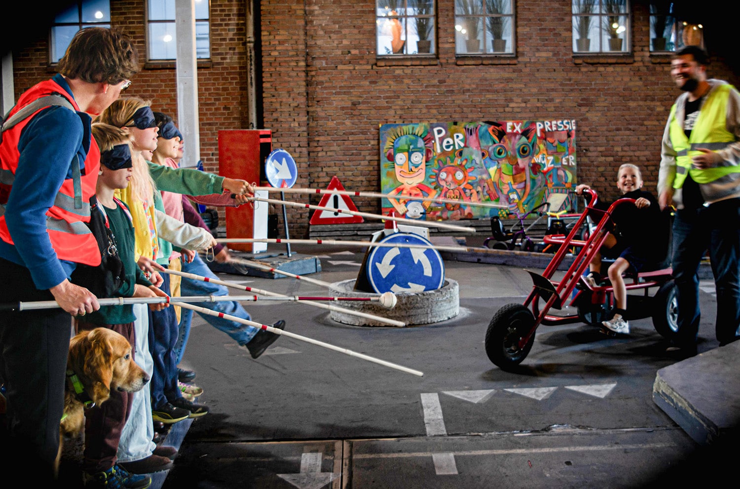 Zeven kinderen met een witte stok en blinddoek steken de weg over in de verkeerstuin in Amersfoort. Een jongen op een skelter wacht geduldig met een grote glimlach.